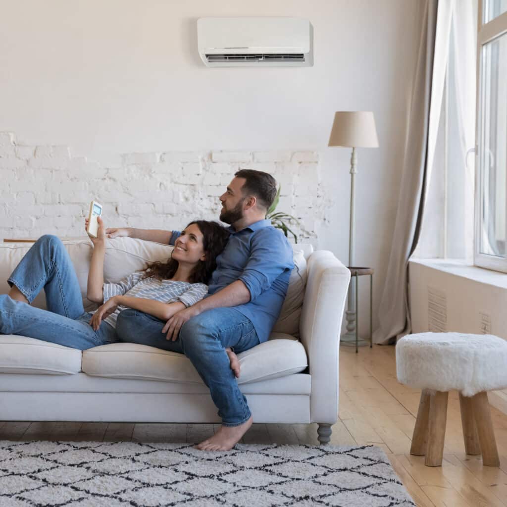 Climatisation maison : couple détendu et confort optimal. Couple détendu sur un canapé blanc, la femme règle la climatisation murale avec une télécommande dans un salon lumineux.