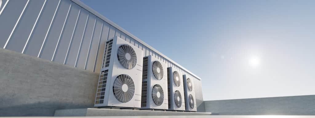 Unités de climatisation extérieures blanches alignées sur une structure moderne en béton et bardage métallique, sous un ciel bleu ensoleillé.