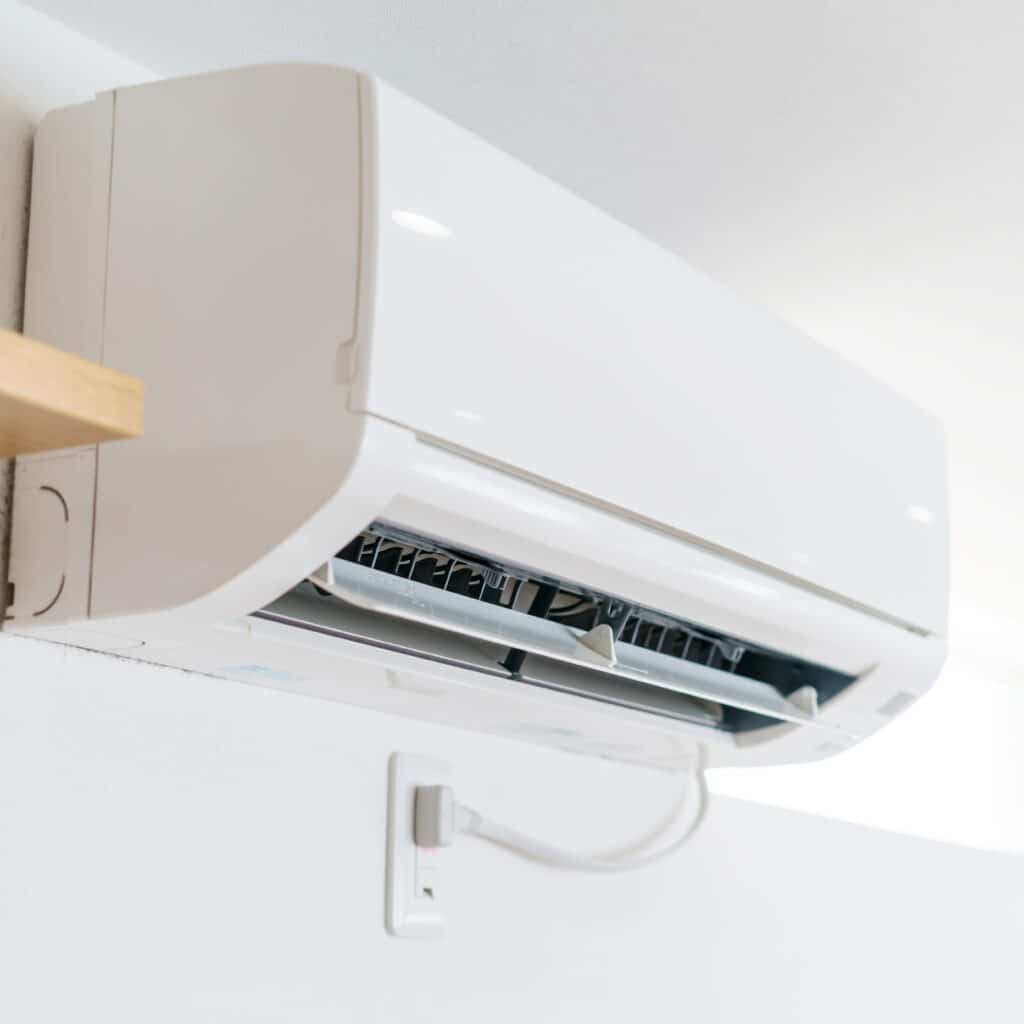 Climatiseur mural blanc moderne avec volets ouverts, branché sur une prise électrique, fixé sur un mur texturé lumineux.