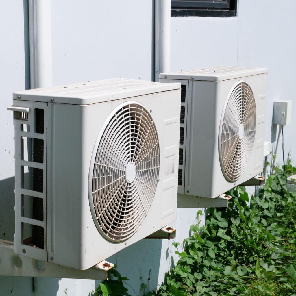 Climatiseurs Extérieurs: Installation sur Mur et Entretien Deux unités extérieures de climatisation blanches sur mur clair, avec ventilateurs visibles, et feuillage vert grimpant en dessous.