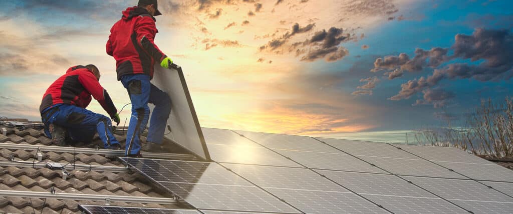 Deux installateurs posent des panneaux solaires sur un toit de tuiles. L'un connecte des câbles, l'autre place un panneau sous un ciel de coucher de soleil.
